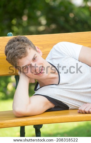 A young male model is smiling at the camera and laying relaxedly on a beautiful wooden bench in a green garden resting his head on his arm