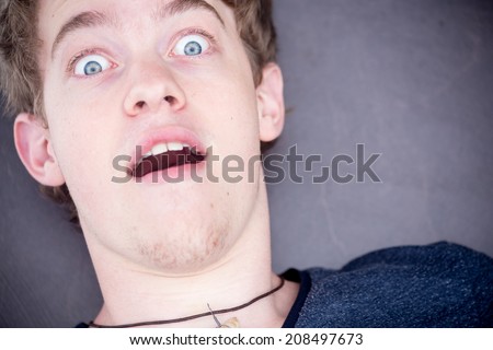 A young blonde male model is very afraid and looks directly into the camera, raising his eyebrows with a nice modern grey background