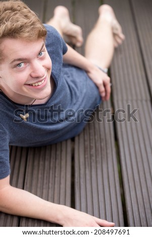 A young blonde male model is lying on the ground, smiling into the camera with his head a bit down