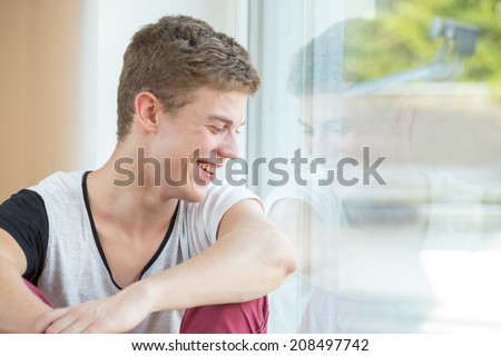 A young male model is laughing down while sitting relaxed next to a window