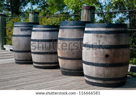 Four wooden barrels along a wooden deck.  Barrels have black metal straps around them.  They are similar to whiskey barrels, but are used for gun powder.