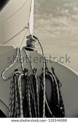Several lines or ropes coiled and tied to a sail boat.  There is one line that is wrapped around the winch and sail is set and visible in the background