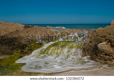 ocean wave rolling over rocks on a beach.  rocks are covered in gree algae and white foam in visible on the water.  copy space in the clear blue sky