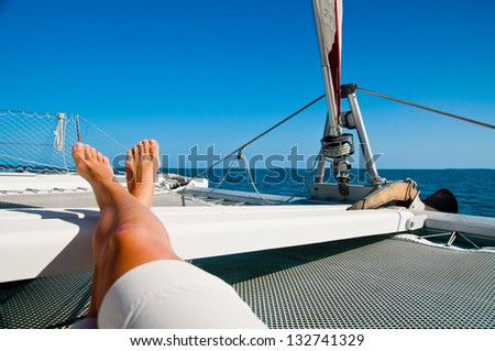 Woman Lounging On A Catamaran Sailboat Trampoline With Her Feet Propped Up And Crossed. Calm Blue Ocean And Cloudless Blue Sky Are In The Background. Copy Space Available