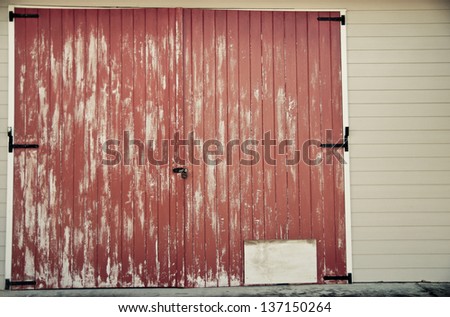 Faded red barn door with a blank white square in the bottom corner for copy space.  Barn doors have metal hinges and are part of a white or cream colored building