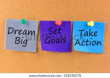 The words Dream Big, Set Goals, Take Action written on sticky colored paper over cork board