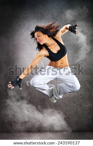 Young beautiful athletic woman jumping dancing modern style dance hip-hop on studio on wall background with smoke