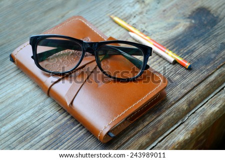 An old notebook in leather cover with pencils and glasses on wooden table
