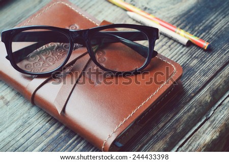 An old notebook in leather cover with pencils and glasses on wooden table