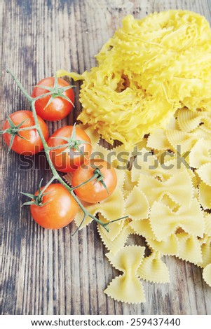 Bunch of small red cherry tomatoes with raw pasta Reginette (Mafaldine) and butterfly shaped pasta farfalle on wooden table
