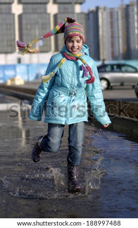 girl in blue jacket and colorful hat is running through spring puddles