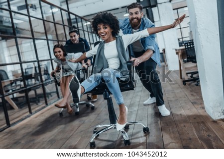 Office fun. Four young cheerful business people in smart casual wear having fun while racing on office chairs and smiling
