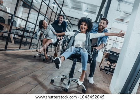 Work hard play hard! Four young cheerful business people in smart casual wear having fun while racing on office chairs and smiling
