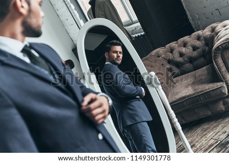 Perfect look. Reflection of handsome young man in full suit adjusting his jacket while standing in front of the mirror indoors