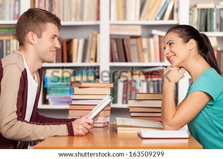 Friends in library. Cheerful young man and woman sitting at the library desk and smiling to each other