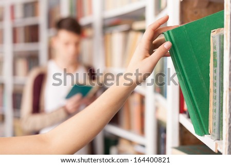 She has got the book she needs. Someone taking a book from the book shelf while young man reading on the background
