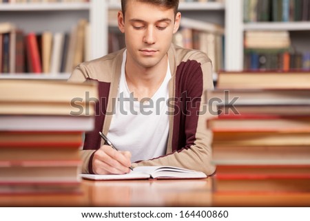 Doing his research in library. Confident young man writing something in his note pad while sitting at the library desk