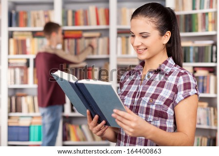 Students in library. Beautiful young woman reading a book while man choosing book from the book shelf on the background