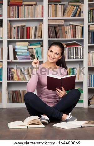Getting ready for her final exam. Beautiful young female student holding book and smiling while sitting on the floor at the library