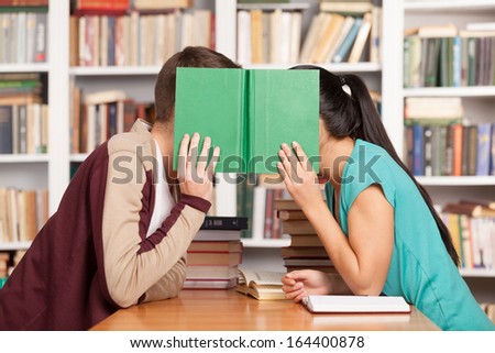 Library romance. Young man and woman sitting close to each other at the library desk and hiding their faces behind a book