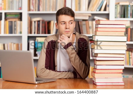 Hard choice. Confused young man sitting at the library desk with a book stack and laptop on it