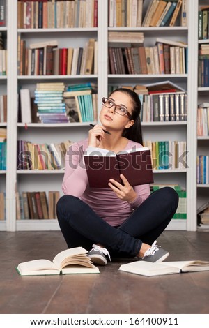 Preparing to the exams. Thoughtful young woman holding a book and looking away while sitting on the floor at the library