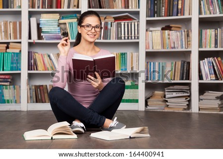 She likes studying. Cheerful young female student holding books and smiling while sitting on the floor at the library