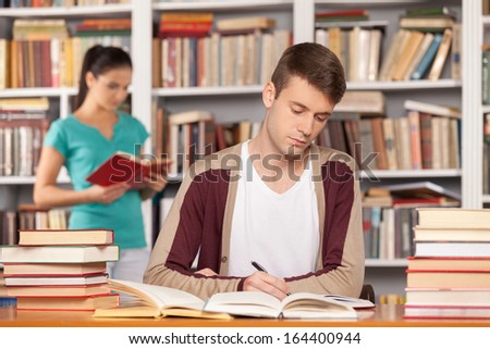Preparing to the exams. Thoughtful young man sitting at the library desk and writing something in his note pad while young woman reading a book on the background