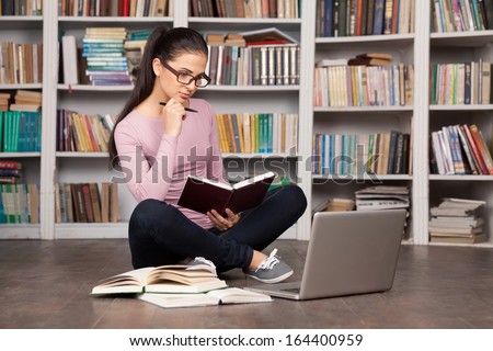 Preparing to the exams. Thoughtful young woman holding a book and looking at the laptop while sitting on the floor at the library