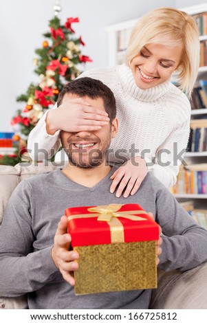 Surprise! Handsome young man sitting on the couch and holding a gift box while her girlfriend standing behind him and covering his eyes with hands