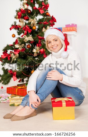 It is Christmas! Cheerful young woman in Christmas hat sitting near the Christmas tree and smiling