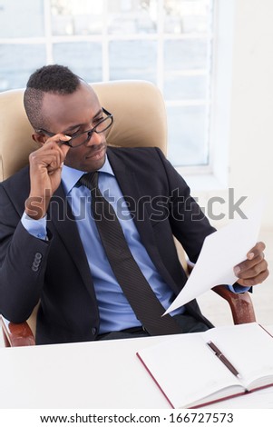 Businessman at work. Top view of confident young African businessman looking at the paper and adjusting his glasses while sitting at his working place