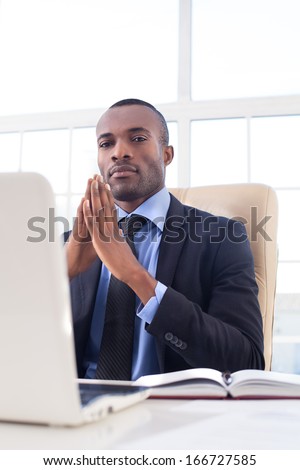 Confident in his power. Confident young African businessman keeping his hands clasped and looking at camera while sitting at his working place