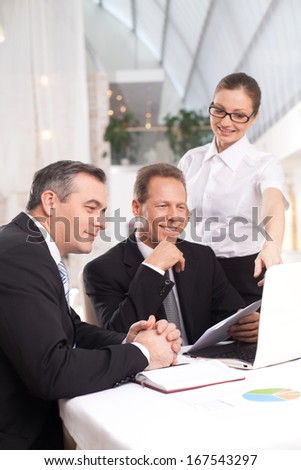 It was a good deal. Two cheerful men in formalwear sitting at the table while woman in white shirt standing close to them and pointing computer monitor