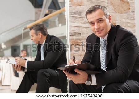 Businessman making notes. Confident mature man in formalwear making notes in his note pad and smiling at camera while sitting on staircase