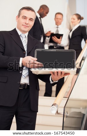 Businessman with laptop. Cheerful mature man in formalwear standing on staircase and pointing laptop while three people talking on background