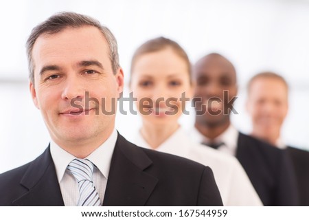 Confident In His Team. Confident Mature Man In Formalwear Looking At Camera And Smiling While His Colleagues Standing In Line Behind Him