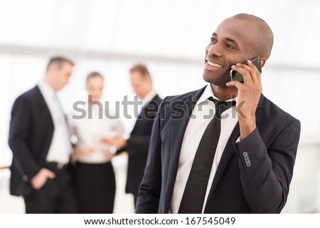 Businessman on the phone. Cheerful young African man in formalwear talking on the mobile phone and smiling while his colleagues standing on background