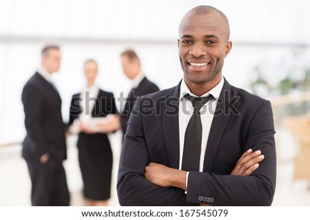 Confident businessman. Cheerful young African man in formalwear keeping arms crossed and smiling while his colleagues standing on background