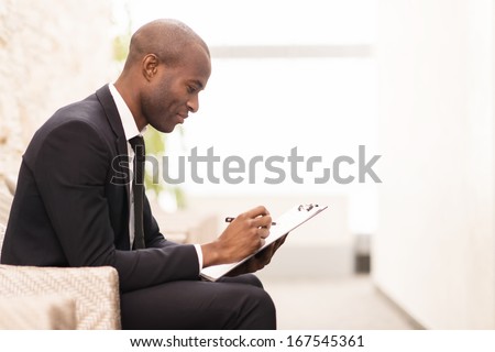 Making notes. Side view of cheerful young African businessman writing something in his note pad and smiling while sitting on the chair