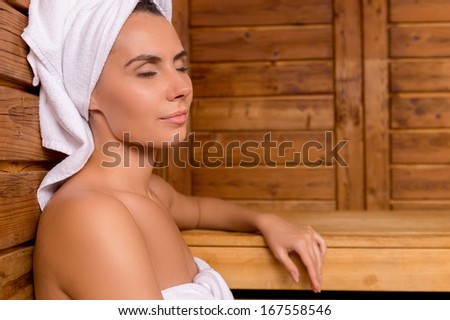 Beauty in sauna. Attractive young woman wrapped in towel relaxing in sauna and keeping eyes closed