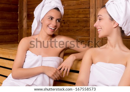 Girls in sauna. Two attractive women wrapped in towel talking to each other and smiling while relaxing in sauna