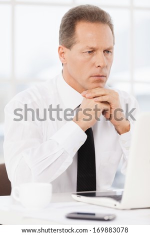 Thoughtful businessman. Thoughtful mature man in shirt and tie holding hand on chin and looking at laptop while sitting at his working place