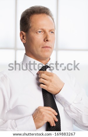 Morning dress up. Confident senior man in white shirt adjusting his necktie and looking away