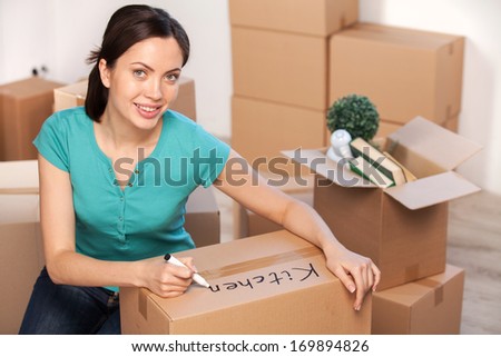 Writing on a carton box. Beautiful young woman marking a cardboard box and smiling at camera while more boxes laying on background