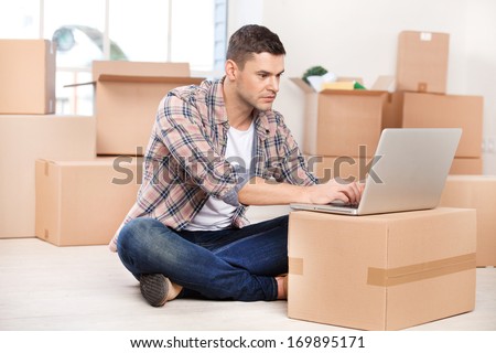 Working in brand new house. Concentrated young man sitting on the floor and working on laptop while cardboard boxes laying on the background