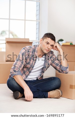 So much work ahead. Depressed young man sitting on the floor and leaning at the carton box while more cardboard boxes laying on the background