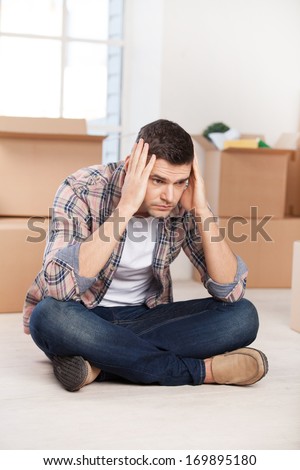 So many boxes to unpack. Depressed young man sitting on the floor and holding head in hands while cardboard boxes laying on the background