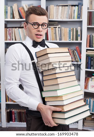 Carrying a book stack. Shocked young man in shirt and bow tie carrying a heavy book stack and looking at camera