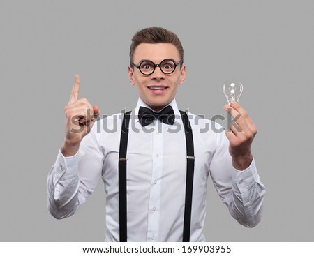 He has got an idea. Surprised young man in bow tie and suspenders holding a light bulb and gesturing while standing against grey background